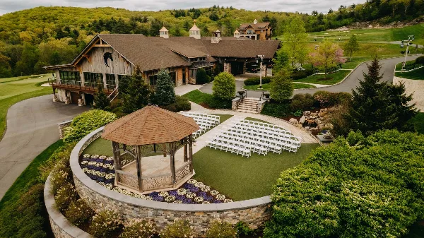Aerial view of a rustic lodge and outdoor wedding ceremony area with a gazebo and rows of white chairs.