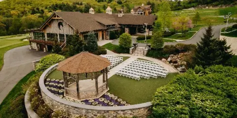 Aerial view of a rustic lodge and outdoor wedding ceremony area with a gazebo and rows of white chairs.
