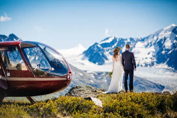 A bride and groom standing on a rocky ledge overlooking a glacier with a red helicopter parked nearby.