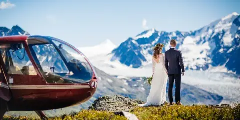 A bride and groom standing on a rocky ledge overlooking a glacier with a red helicopter parked nearby.