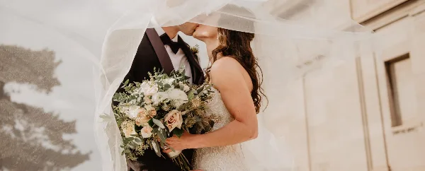 A bride in a white lace gown holding a large bouquet of white and blush roses with eucalyptus greenery while kissing a groom under a veil.