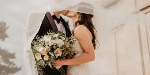 A bride in a white lace gown holding a large bouquet of white and blush roses with eucalyptus greenery while kissing a groom under a veil.