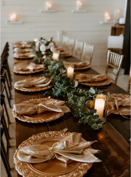 Long wooden wedding reception table set with gold charger plates, napkins, candles, and a greenery centerpiece, with chairs along both sides.