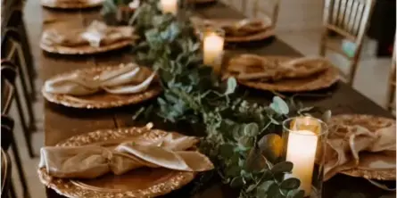 Long wooden wedding reception table set with gold charger plates, napkins, candles, and a greenery centerpiece, with chairs along both sides.