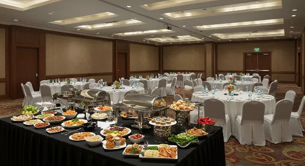 A catering buffet table with chafing dishes and assorted food platters set up in a banquet hall with round tables.