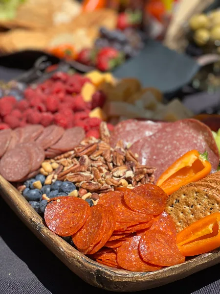 A close-up of a catering charcuterie board featuring sliced salami, cured meats, pecans, blueberries, raspberries, and crackers.