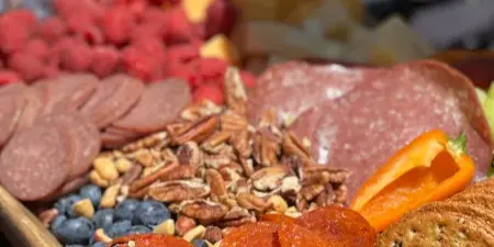 A close-up of a catering charcuterie board featuring sliced salami, cured meats, pecans, blueberries, raspberries, and crackers.