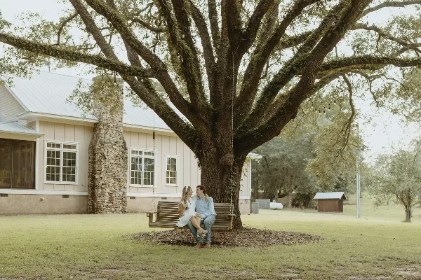 A couple sitting on a wooden swing attached to a large oak tree in front of a white barn with a stone chimney.