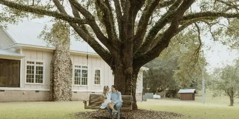 A couple sitting on a wooden swing attached to a large oak tree in front of a white barn with a stone chimney.