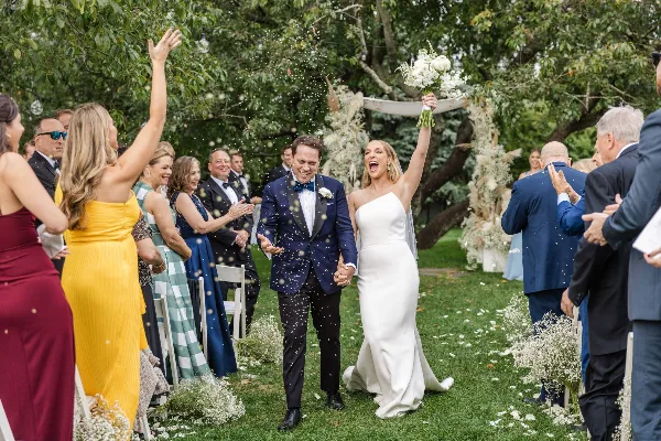 A bride and groom walking down an outdoor wedding aisle with guests and confetti.
