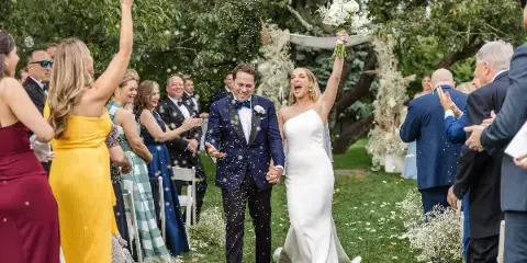 A bride and groom walking down an outdoor wedding aisle with guests and confetti.