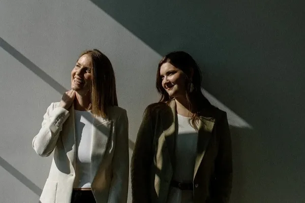 Two smiling women in blazers standing against a sunlit wall, representing the Meadowlark Films wedding videography team.