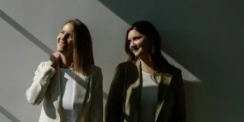 Two smiling women in blazers standing against a sunlit wall, representing the Meadowlark Films wedding videography team.