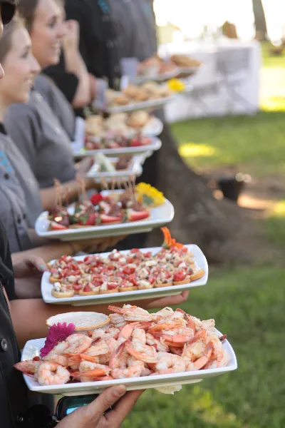 Catering staff serving platters of shrimp, bruschetta, and other appetizers at an outdoor event.
