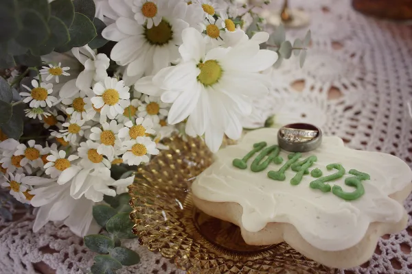 A frosted sugar cookie with a wedding ring and white daisies on a lace tablecloth.
