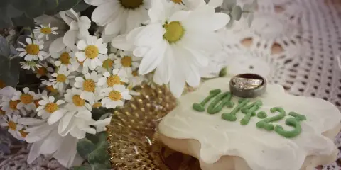 A frosted sugar cookie with a wedding ring and white daisies on a lace tablecloth.