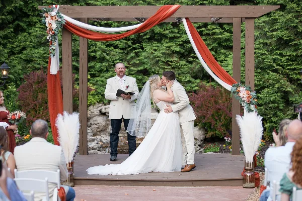 A bride and groom sharing a kiss under a wooden pergola decorated with red and white fabric during an outdoor wedding ceremony, with an officiant and guests present.