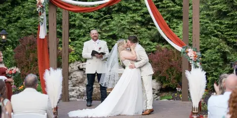 A bride and groom sharing a kiss under a wooden pergola decorated with red and white fabric during an outdoor wedding ceremony, with an officiant and guests present.