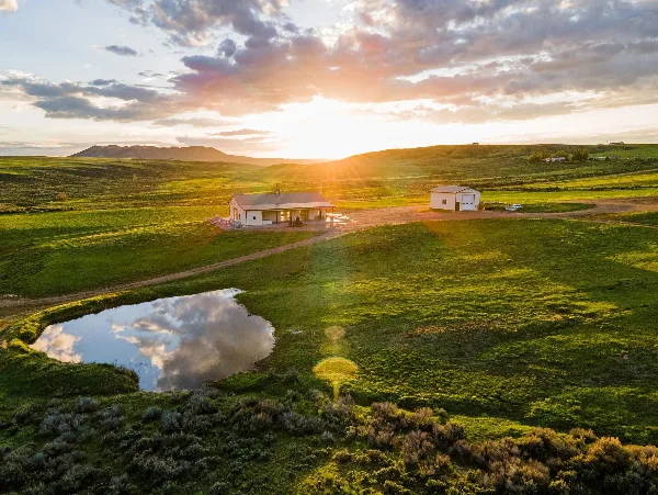 Aerial view of a rural ranch venue with an event building, guest cabin, pond, and rolling green fields at sunset.