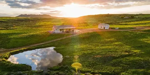 Aerial view of a rural ranch venue with an event building, guest cabin, pond, and rolling green fields at sunset.