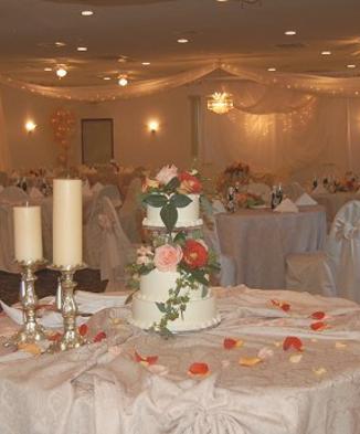 A wedding reception venue featuring a decorated three-tier wedding cake with roses, candles, and set dining tables in the background.