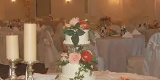 A wedding reception venue featuring a decorated three-tier wedding cake with roses, candles, and set dining tables in the background.