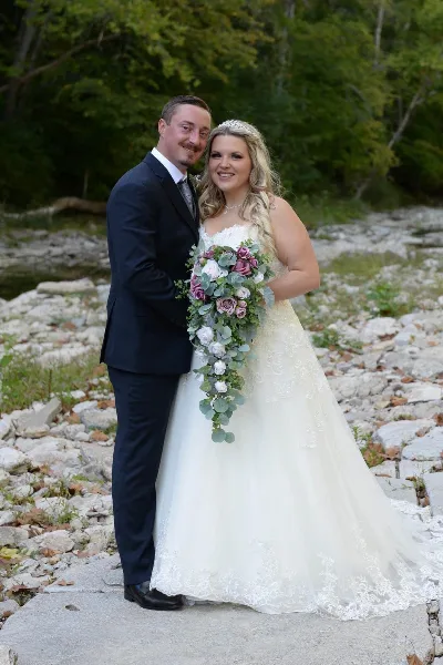 A bride and groom posing together outdoors near a rocky creek bed with a forest background.
