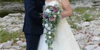A bride and groom posing together outdoors near a rocky creek bed with a forest background.