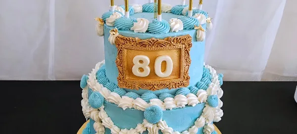 A two-tier blue and white custom celebration cake decorated with piped frosting, gold accents, and the number 80.