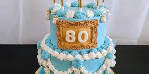 A two-tier blue and white custom celebration cake decorated with piped frosting, gold accents, and the number 80.