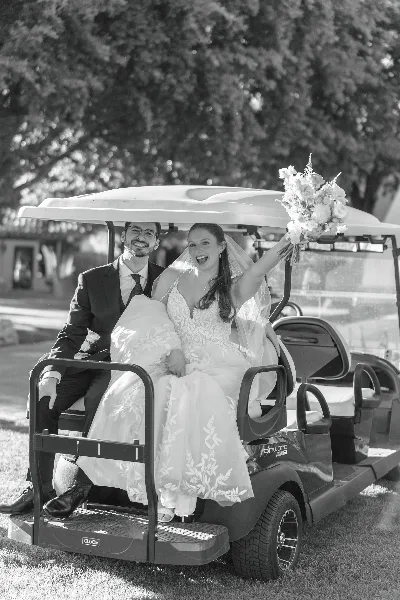 A bride and groom smiling while sitting on a golf cart, with the bride holding a bouquet.