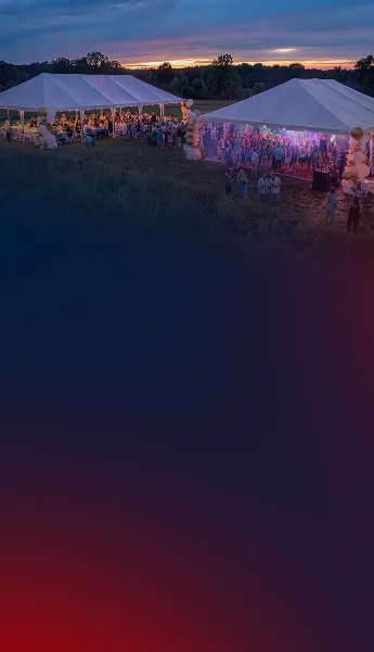 Two large white event tents set up in a grassy field at dusk, filled with guests and decorated with balloons.