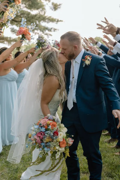 A bride and groom share a kiss outdoors while surrounded by guests holding bouquets.