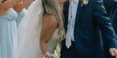 A bride and groom share a kiss outdoors while surrounded by guests holding bouquets.