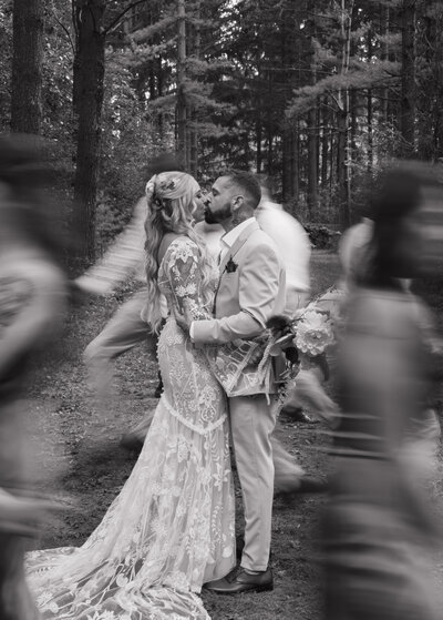 A black and white photograph of a bride and groom kissing in a forest, surrounded by blurred figures of dancing guests.