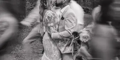 A black and white photograph of a bride and groom kissing in a forest, surrounded by blurred figures of dancing guests.