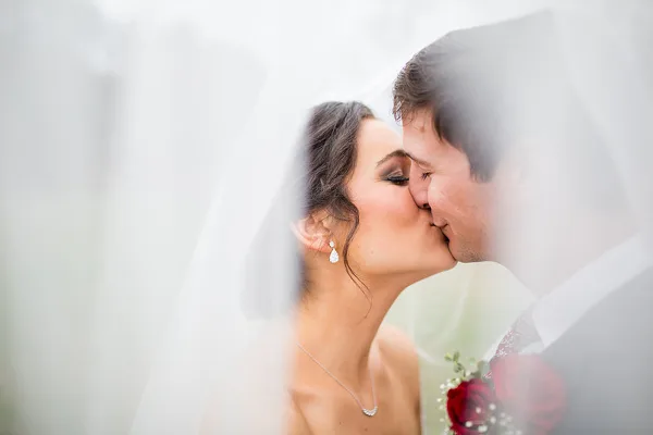 A bride and groom sharing a kiss, with the bride wearing a veil and holding a red rose bouquet.