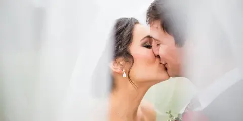 A bride and groom sharing a kiss, with the bride wearing a veil and holding a red rose bouquet.