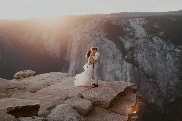 A bride and groom in wedding attire embracing on a rocky cliff edge with a mountainous valley in the background.