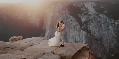 A bride and groom in wedding attire embracing on a rocky cliff edge with a mountainous valley in the background.