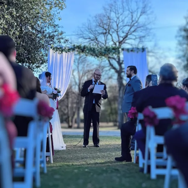 Outdoor wedding ceremony with bride and groom at an arch, officiant speaking into a microphone, and guests seated in white chairs.