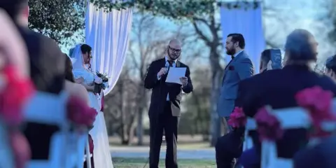 Outdoor wedding ceremony with bride and groom at an arch, officiant speaking into a microphone, and guests seated in white chairs.