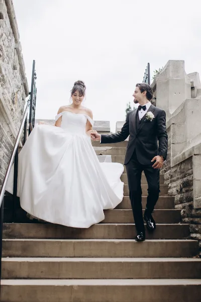 A bride in a white gown and a groom in a black tuxedo walking down stone steps while holding hands.