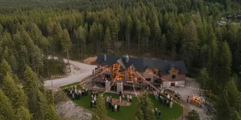 Aerial view of a large mountain lodge at dusk with a wedding ceremony taking place on the lawn surrounded by string lights and forest.
