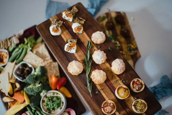 A wooden serving board featuring assorted appetizers including cheese-topped crackers, powdered sugar-dusted pastries, and small tartlets, alongside a side platter of fresh vegetables and dips.