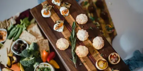 A wooden serving board featuring assorted appetizers including cheese-topped crackers, powdered sugar-dusted pastries, and small tartlets, alongside a side platter of fresh vegetables and dips.