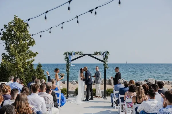 A wedding ceremony in progress on a beachfront patio with a view of the lake and a decorated arch.