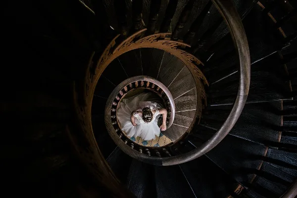 A bride in a white wedding dress standing on a dark wooden spiral staircase, viewed from above.