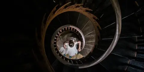 A bride in a white wedding dress standing on a dark wooden spiral staircase, viewed from above.