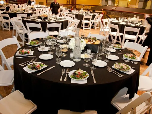 A round banquet table set with black linens, white plates, silverware, and a salad dish in a large event venue.